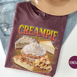 May include: A maroon t-shirt with the text "CREAMPIE" in yellow and orange letters. The shirt features a graphic of various pies, including a cream pie, a coconut cream pie, a cheesecake, and a caramel pie.