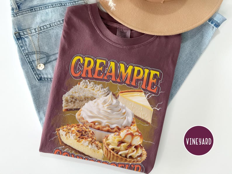 May include: A maroon t-shirt with the text "CREAMPIE" in yellow and orange letters. The shirt features a graphic of various pies, including a cream pie, a coconut cream pie, a cheesecake, and a caramel pie.