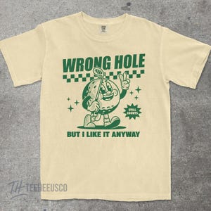 May include: Cream-colored t-shirt with a green graphic of a golf ball character holding a golf club and giving a peace sign. Text reads "WRONG HOLE" and "BUT I LIKE IT ANYWAY". The shirt has a vintage, cartoon-like design.