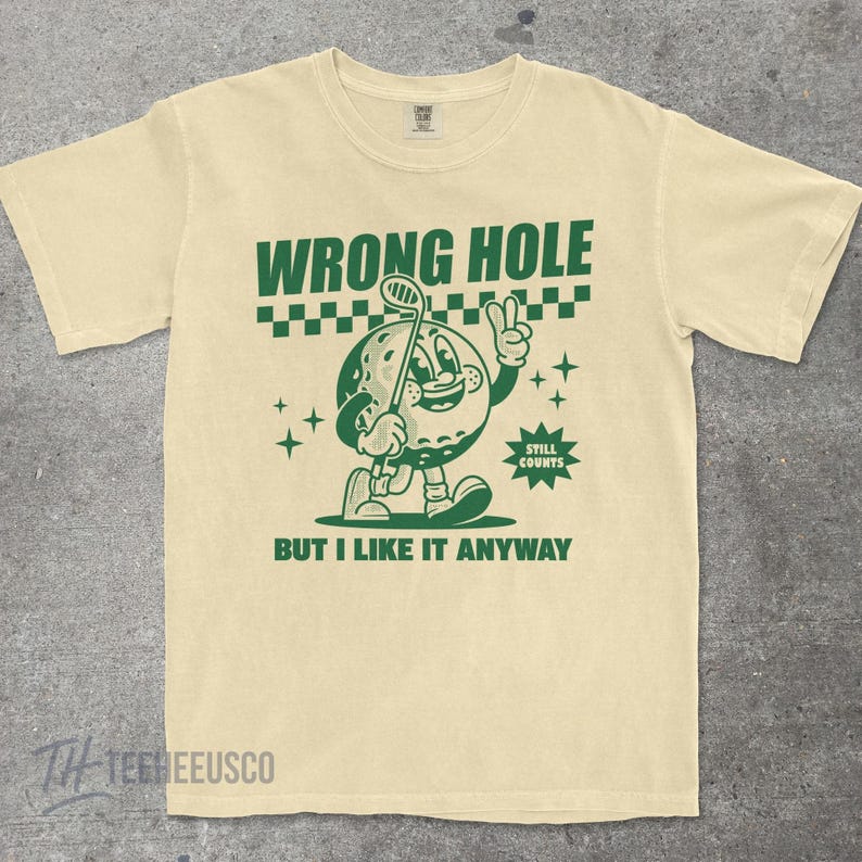 May include: Cream-colored t-shirt with a green graphic of a golf ball character holding a golf club and giving a peace sign. Text reads "WRONG HOLE" and "BUT I LIKE IT ANYWAY". The shirt has a vintage, cartoon-like design.