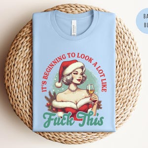 Pu&ograve; includere: T-shirt azzurra con una grafica di una donna con un cappello di Babbo Natale che tiene in mano un bicchiere di vino. Il testo sulla maglietta recita "It's beginning to look a lot like Fuck This."