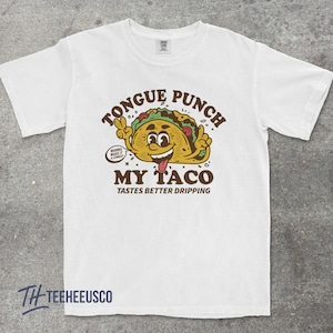 May include: White t-shirt with a cartoon taco graphic. The taco has a smiling face and is making a peace sign with its hands. Text on the shirt reads "TONGUE PUNCH MY TACO TASTES BETTER DRIPPING" and "WANNA MAKE IT A COMBO?"