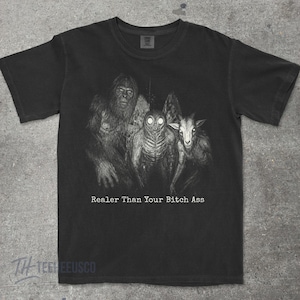 May include: A black short-sleeve t-shirt featuring a graphic print of Bigfoot, the Mothman, and a goat-like creature. Below the creatures, the text reads, "Realer Than Your Bitch Ass" in a white, typewriter-style font. The shirt has a crew neckline.