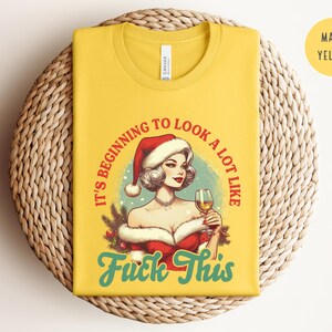 Pu&ograve; includere: T-shirt giallo mais con grafica di una donna con cappello da Babbo Natale che tiene in mano un calice di vino. Il testo sulla maglietta recita "It's beginning to look a lot like Fuck This" in rosso e verde acqua.
