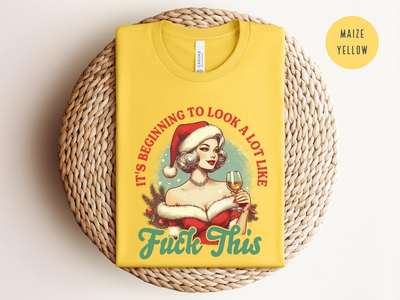 Pu&ograve; includere: T-shirt giallo mais con grafica di una donna con cappello da Babbo Natale che tiene in mano un calice di vino. Il testo sulla maglietta recita "It's beginning to look a lot like Fuck This" in rosso e verde acqua.