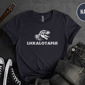Może przedstawiać: Koszulka w kolorze granatowym z białym nadrukiem szkieletu dinozaura i tekstem "LICKALOTAPUS".