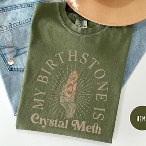 May include: A green t-shirt with the text "My Birthstone Is Crystal Meth" in a circle with a pink crystal graphic inside. The shirt is made of hemp.