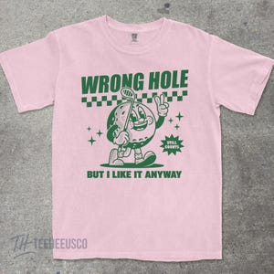May include: Pink t-shirt with a green cartoon golf ball character holding a golf club and giving a peace sign. The shirt has the text "WRONG HOLE" above the graphic and "BUT I LIKE IT ANYWAY" below.