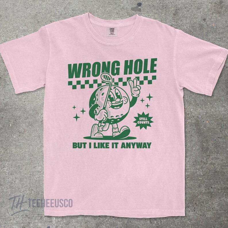 May include: Pink t-shirt with a green cartoon golf ball character holding a golf club and giving a peace sign. The shirt has the text "WRONG HOLE" above the graphic and "BUT I LIKE IT ANYWAY" below.