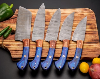 May include: A set of five Damascus steel kitchen knives with blue and wood handles, arranged on a wooden cutting board. The knives have a unique pattern on the blades. Green bell pepper, chili peppers, tomato, onion, and lemons are also in the image.