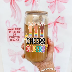 May include: A clear glass tumbler with a bamboo lid and a glass straw, filled with a brown beverage. The tumbler features a colorful sticker with the text "CHEERS QUEERS" and peace signs with rainbow armbands. The background has pink bow illustrations.