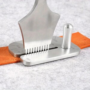 May include: A silver-colored metal leathercraft corner punch tool with a vertical handle and a flat base featuring etched markings R5, R15, R20, and 40. The tool is positioned over a strip of orange leather, showing a series of small teeth.