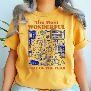 Op de afbeelding: Mosterdgeel T-shirt met een afbeelding van spoken die boeken lezen in een spookachtige boekwinkel. De tekst luidt "The Most Wonderful Time of the Year" en "Haunted Bookstore Spooky Sale."
