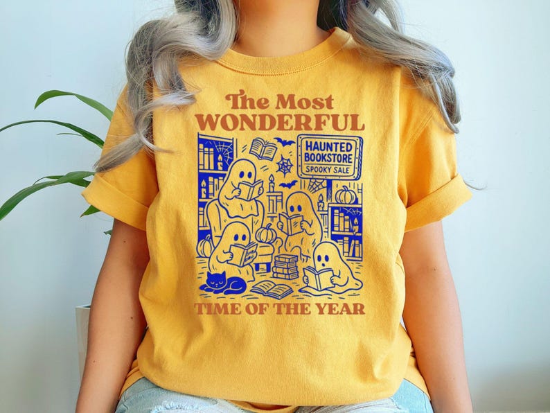 Op de afbeelding: Mosterdgeel T-shirt met een afbeelding van spoken die boeken lezen in een spookachtige boekwinkel. De tekst luidt "The Most Wonderful Time of the Year" en "Haunted Bookstore Spooky Sale."