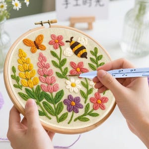 Op de afbeelding: Een ronde houten borduurring met een beige stoffen paneel met een handgeborduurd ontwerp van een gele vlinder, een hommel en diverse kleurrijke bloemen, waaronder gele en roze lupinen, madeliefjes en paarse bloesems.