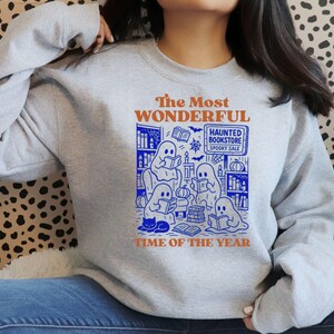 Op de afbeelding: Een lichtgrijze sweatshirt met een blauwe en oranje afbeelding. De afbeelding toont spoken die boeken lezen in een boekhandel, met de tekst "The Most Wonderful Time of the Year" en "Haunted Bookstore Spooky Sale".
