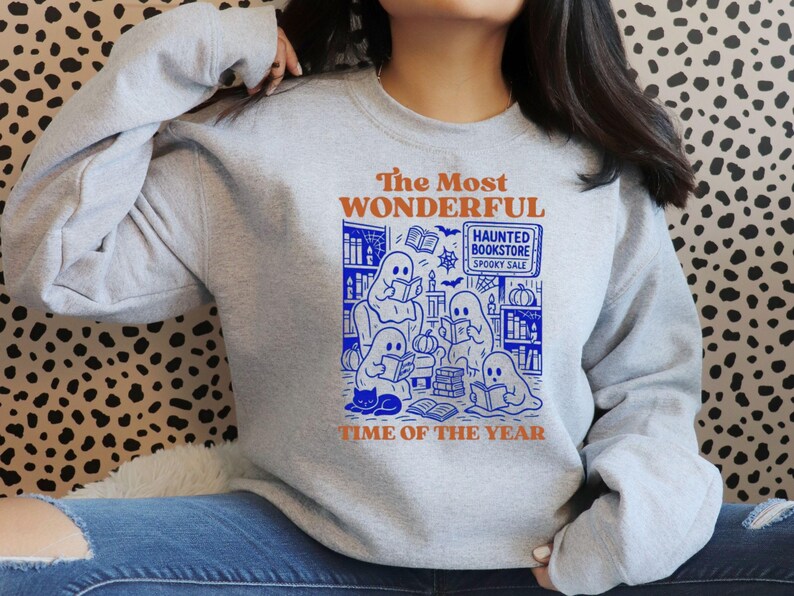 Op de afbeelding: Een lichtgrijze sweatshirt met een blauwe en oranje afbeelding. De afbeelding toont spoken die boeken lezen in een boekhandel, met de tekst "The Most Wonderful Time of the Year" en "Haunted Bookstore Spooky Sale".