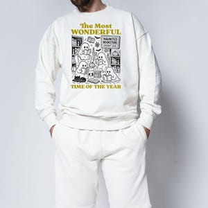 Op de afbeelding: Cr&egrave;mekleurige sweatshirt- en shortset. Het sweatshirt heeft een zwart-witte afbeelding van spoken die boeken lezen in een boekhandel, met de tekst "The Most Wonderful Time of the Year".