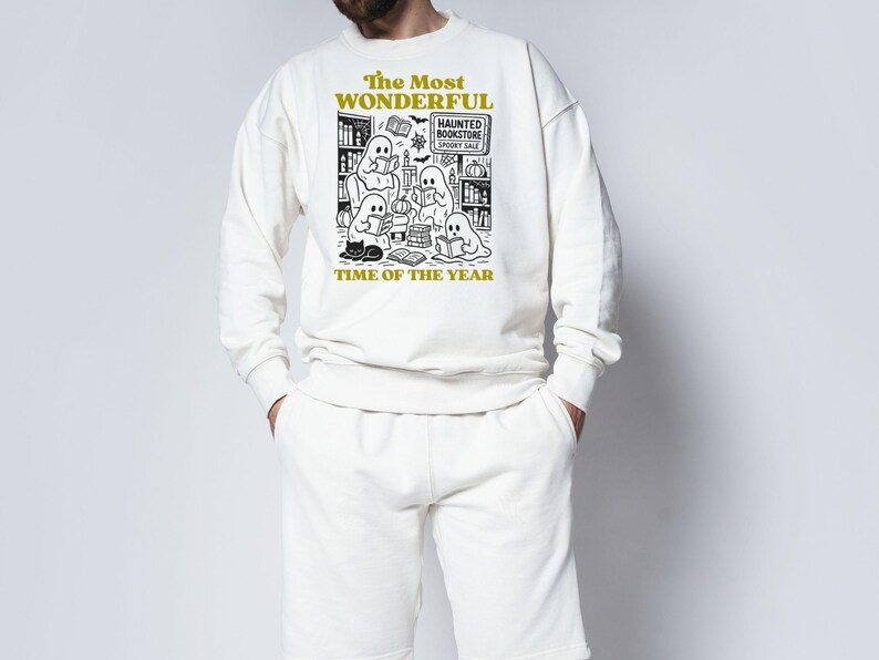 Op de afbeelding: Cr&egrave;mekleurige sweatshirt- en shortset. Het sweatshirt heeft een zwart-witte afbeelding van spoken die boeken lezen in een boekhandel, met de tekst "The Most Wonderful Time of the Year".