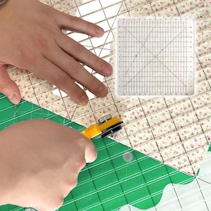 Puede incluir: Una regla de acolchado cuadrada transparente con marcas de cuadrícula negras y líneas diagonales, colocada sobre tela floral en una base de corte verde. Un cortador rotativo amarillo con hoja circular corta la tela junto a la regla.