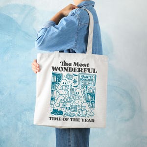 Op de afbeelding: Cr&egrave;mekleurige draagtas met een teal illustratie van geesten die in een boekhandel lezen. De tekst op de tas luidt "The Most Wonderful Time of the Year" en "Haunted Bookstore Spooky Sale".