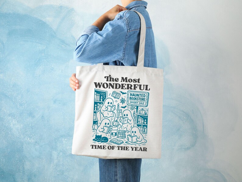 Op de afbeelding: Cr&egrave;mekleurige draagtas met een teal illustratie van geesten die in een boekhandel lezen. De tekst op de tas luidt "The Most Wonderful Time of the Year" en "Haunted Bookstore Spooky Sale".