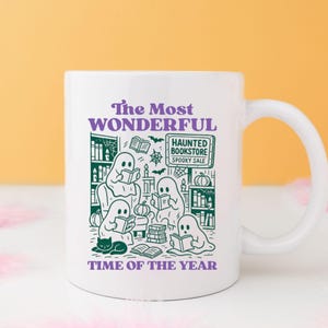 Op de afbeelding: Witte keramische mok met een Halloween-thema. De mok heeft groene illustraties van spoken die boeken lezen, een zwarte kat en de tekst "The Most Wonderful Time of the Year". Bevat de tekst "Haunted Bookstore Spooky Sale".