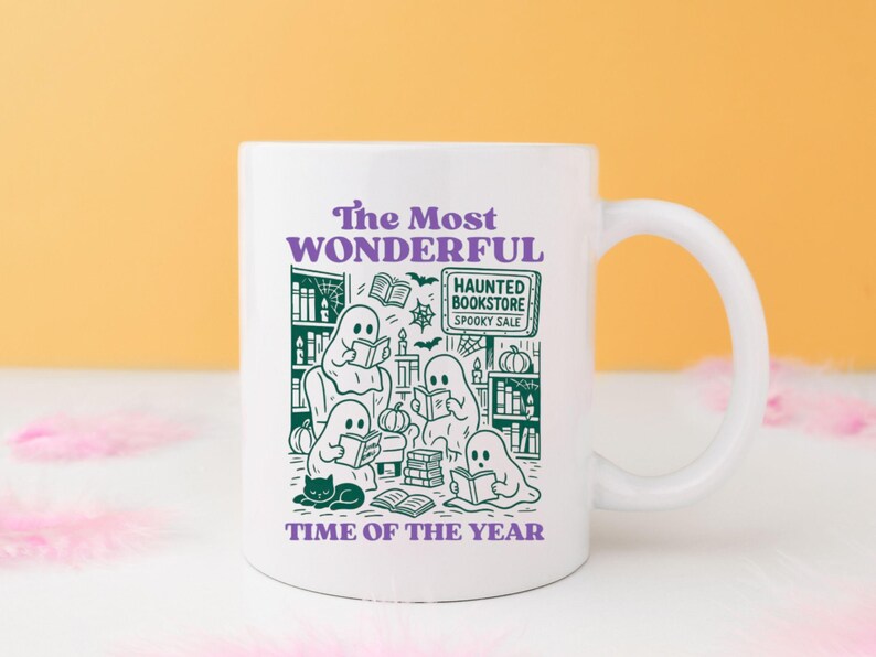 Op de afbeelding: Witte keramische mok met een Halloween-thema. De mok heeft groene illustraties van spoken die boeken lezen, een zwarte kat en de tekst "The Most Wonderful Time of the Year". Bevat de tekst "Haunted Bookstore Spooky Sale".