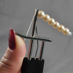 May include: A silver-toned metal bead stringing tool with a black handle, featuring a thin needle-like tip threaded with white string. A row of six small, round, cream-colored pearls is threaded onto the string, extending outward from the needle tip.