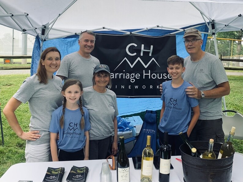 Puede incluir: Un grupo de personas se encuentran de pie frente a una tienda de campa&ntilde;a azul y blanca con un letrero que dice "CH Carriage House Wineworks". Todos llevan camisas con el mismo logotipo. Hay botellas de vino sobre una mesa frente a ellos.