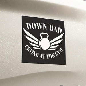 May include: Black square sticker with white text and a kettlebell graphic. The text reads "DOWN BAD CRYING AT THE GYM". The sticker is affixed to the back of a white vehicle, near the taillight.