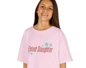 Puede incluir: Una camiseta rosa claro con el texto "Eldest Daughter" en fuente roja, acompañada de tres estrellas turquesas. La camiseta es de estilo casual.