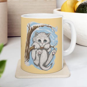 May include: White ceramic mug featuring a whimsical illustration of a gray kitten clinging to a tree branch. The design includes a snowy tree and blue accents, set against a light yellow background. A charming and cozy design.