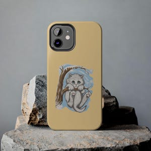 May include: A tan phone case featuring a whimsical illustration of a gray kitten clinging to a tree branch. The artwork includes blue and white accents, creating a charming, artistic design. The phone case is shown on a stone surface.
