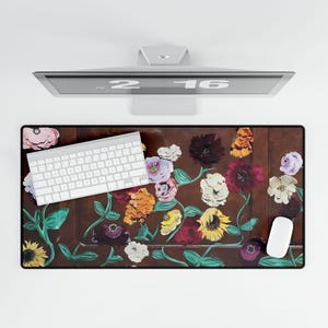May include: A rectangular desk mat with a floral design in shades of yellow, red, purple, and white. A white keyboard and mouse are on the mat. The mat is brown with a black border. A computer monitor is above the mat.