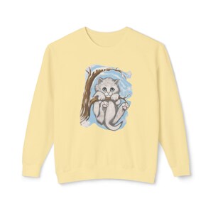 May include: A light yellow sweatshirt featuring a graphic of a gray kitten clinging to a tree branch. The kitten has blue eyes and is set against a blue and white snowy background. The sweatshirt has long sleeves and a crew neck.