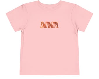 Puede incluir: Camiseta rosa claro para niños pequeños con la palabra "SHOWGIRL" impresa en letras de purpurina naranja y dorada en el pecho. La camiseta de manga corta es de tejido suave.
