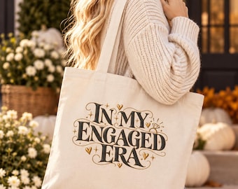 May include: A cream-colored canvas tote bag featuring the black text "IN MY ENGAGED ERA" in a stylized font, accented with small gold hearts, stars, and a diamond ring graphic. The bag has two shoulder straps and a rectangular shape.