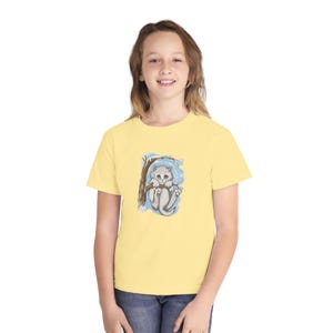Pode incluir: Uma camiseta amarelo claro com uma ilustração em aquarela de um gatinho cinza em um galho de árvore. O gatinho tem olhos azuis e está em um fundo azul e branco. A camiseta é feita de um material macio.