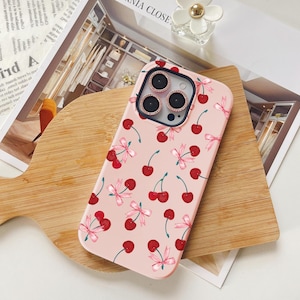 May include: A pink phone case with a cherry and bow pattern. The case has a clear camera lens cover with a decorative ring around each lens.