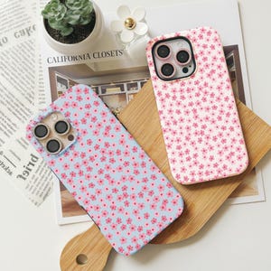 May include: Two phone cases with a floral pattern. The case on the left is light blue with pink flowers. The case on the right is white with pink flowers. Both cases have a camera cutout with three holes.