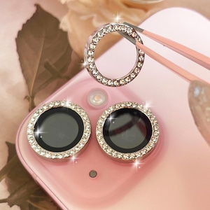 May include: A close-up of a pink phone with two camera lens protectors. Each protector is a silver ring encrusted with small, clear rhinestones. A third, similar ring is held above the phone with pink tweezers. The phone's camera lenses are black.
