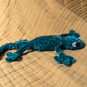 Op de afbeelding: Een tealkleurig pluche hagedisspeelgoed met een fluweelzachte textuur. De hagedis heeft een lange staart, vier poten en een kleine kop met een wit en zwart oog. Het ligt op een bruin, getextureerde stof.