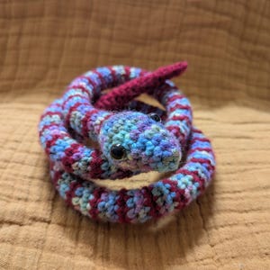 Op de afbeelding: Een opgerolde, handgemaakte gehaakte slang in tinten blauw, paars en rood. De slang heeft zwarte ogen en een rode tong. Het knutselproject staat tegen een getextureerde, bruine achtergrond.