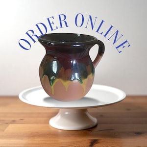 May include: A brown ceramic pitcher with a green and yellow glaze. The pitcher has a handle and is sitting on a white cake stand. The text "ORDER ONLINE" is written in blue letters above the pitcher.