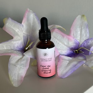May include: A small brown glass bottle with a dropper top. The bottle is labeled "Radiant Rose Apothecary" and "Fear Be Gone Essence Blend". The bottle is sitting on a white surface with two purple and white flowers.