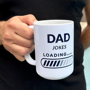 May include: White ceramic mug with the words "DAD JOKES LOADING....." in black text. The mug features a loading bar graphic. The mug is held by a person wearing a black shirt.