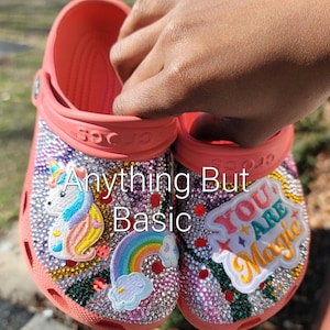 Peut inclure: Une paire de sabots roses décorés de strass et de patchs colorés. Un sabot présente une licorne, un arc-en-ciel et des nuages. L'autre sabot présente le texte "You Are Magic". Le texte "Anything But Basic" est écrit sur le devant des sabots.