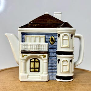 May include: A ceramic teapot shaped like a house, featuring a brown roof, blue siding, and white trim. The teapot has a balcony, bay windows, and a chimney. The handle and spout are white.