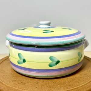 May include: A ceramic lidded serving dish with a yellow base and lid, adorned with green leaf designs. The lid features blue, purple, and pink stripes. The dish has two small handles on either side.